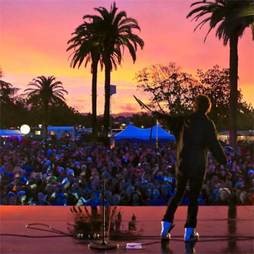 Performer on stage at sunset during 90 Plus Wine Club event in Napa with palm trees and audience.