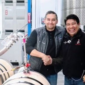Two men shaking hands in a winery or wine production facility with wine barrels and equipment.